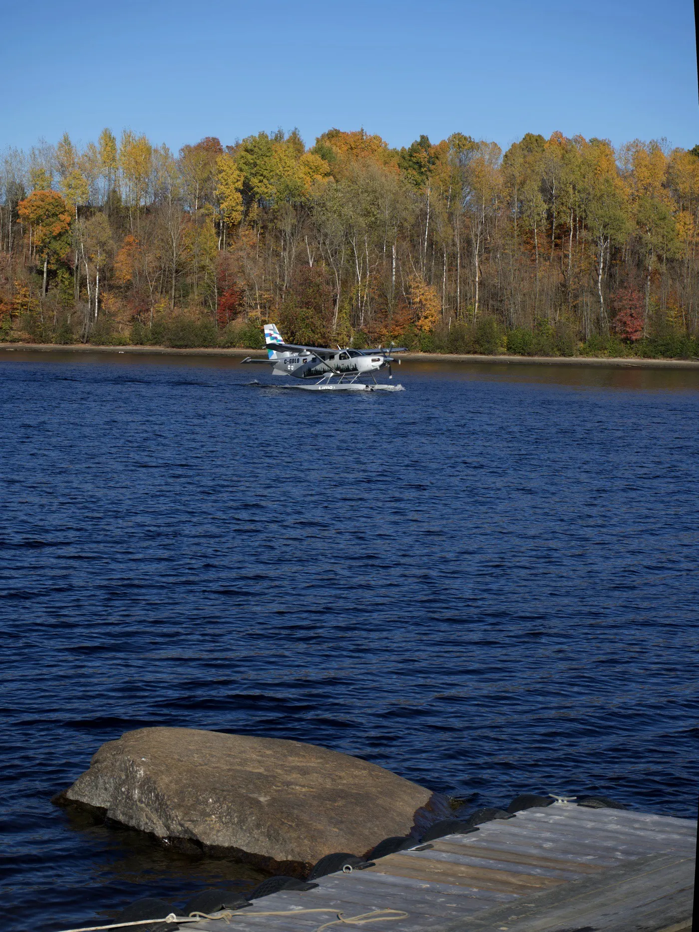 A white seaplane on blue water, with autumn trees behind it and a dock in the foreground in Québec